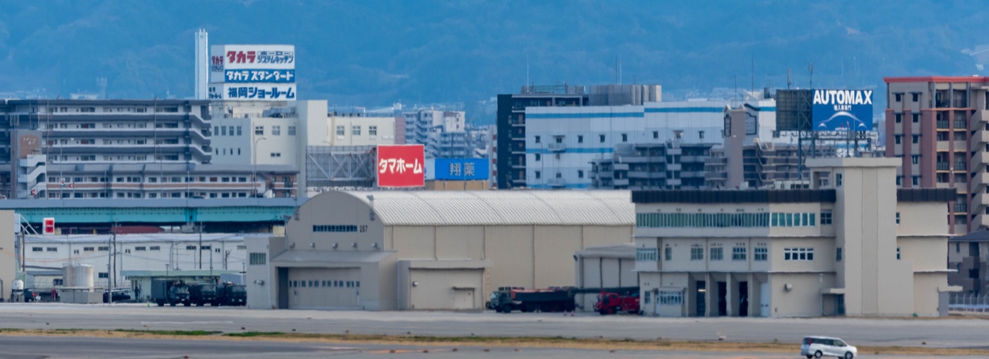 航空自衛隊春日基地の格納庫と、その背後に建ち並ぶマンションや商業施設